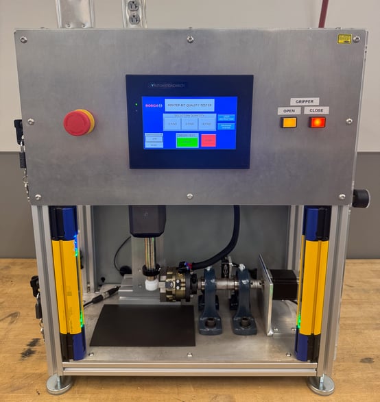 Front view of a metal router bit quality tester machine featuring a digital touch display screen, emergency stop button, physical gripper controls, and yellow safety light curtains. 