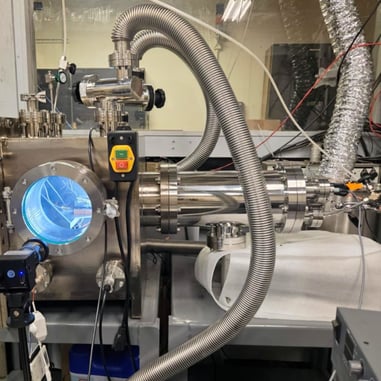 A laboratory photograph of the stainless steel vacuum and high-pressure chamber used at MIT for battery perforation testing. The system features a circular viewport with blue internal lighting, heavy-duty metal hosing, and an external high-speed camera for documenting experiments. 