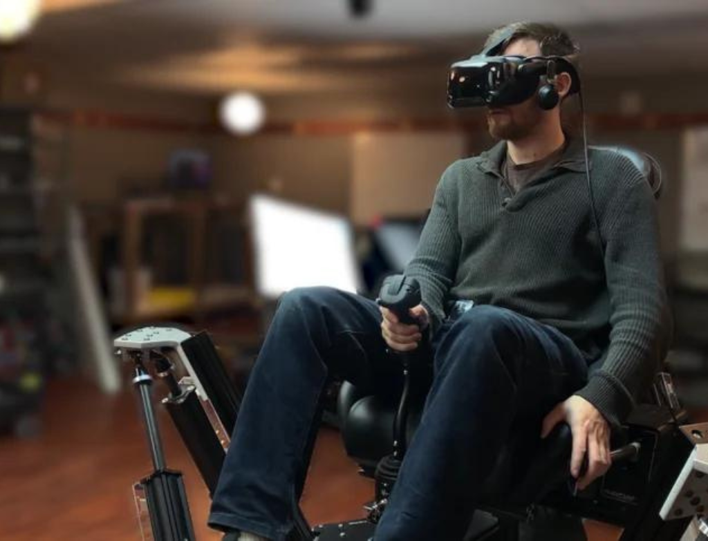 A man sits on a motion platform wearing a VR headset and handling a helicopter cyclic and collective.
