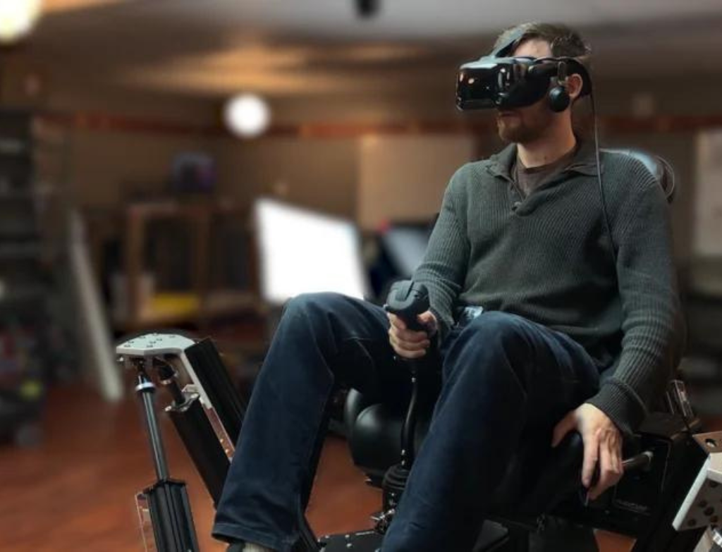 A man sits on a motion platform wearing a VR headset and handling a helicopter cyclic and collective. A man sits on a motion platform wearing a VR headset and handling a helicopter cyclic and collective.