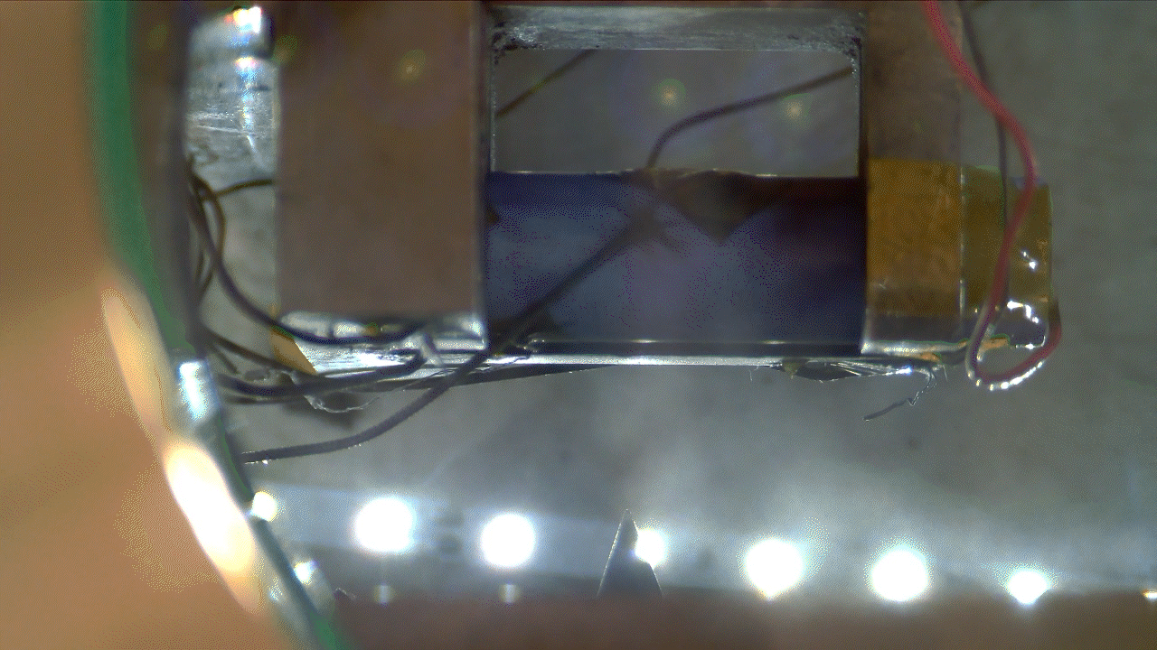 Animated GIF showing a high-speed linear actuator driving a nail into a lithium-ion battery. The perforation causes an immediate spark and a controlled release of gas inside a metallic test chamber. 